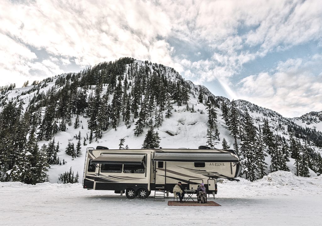 2 people are sitting outside of an RV that is set up in a snowy mountain area